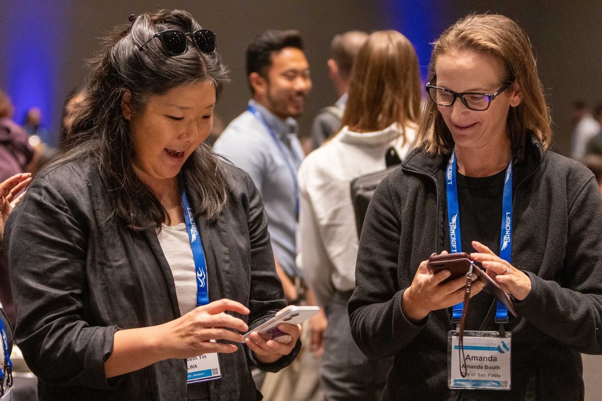 Two attendees laughing over their phones while networking at the CASQA conference — corporate event photography by Michael Pegram