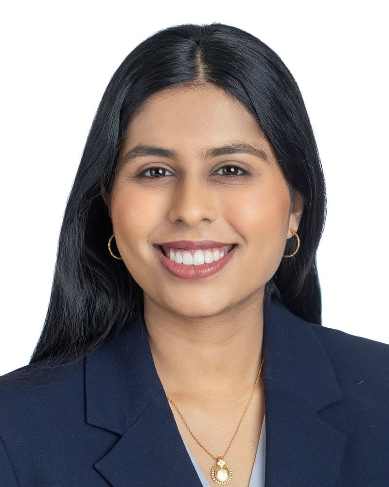 Smiling woman with long black hair in a navy blazer and gold necklace, headshot on white