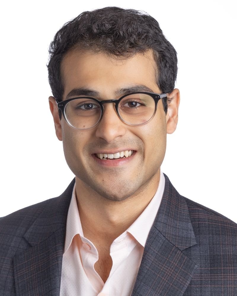 Smiling man in round glasses, grey plaid blazer and pale pink shirt, headshot on white