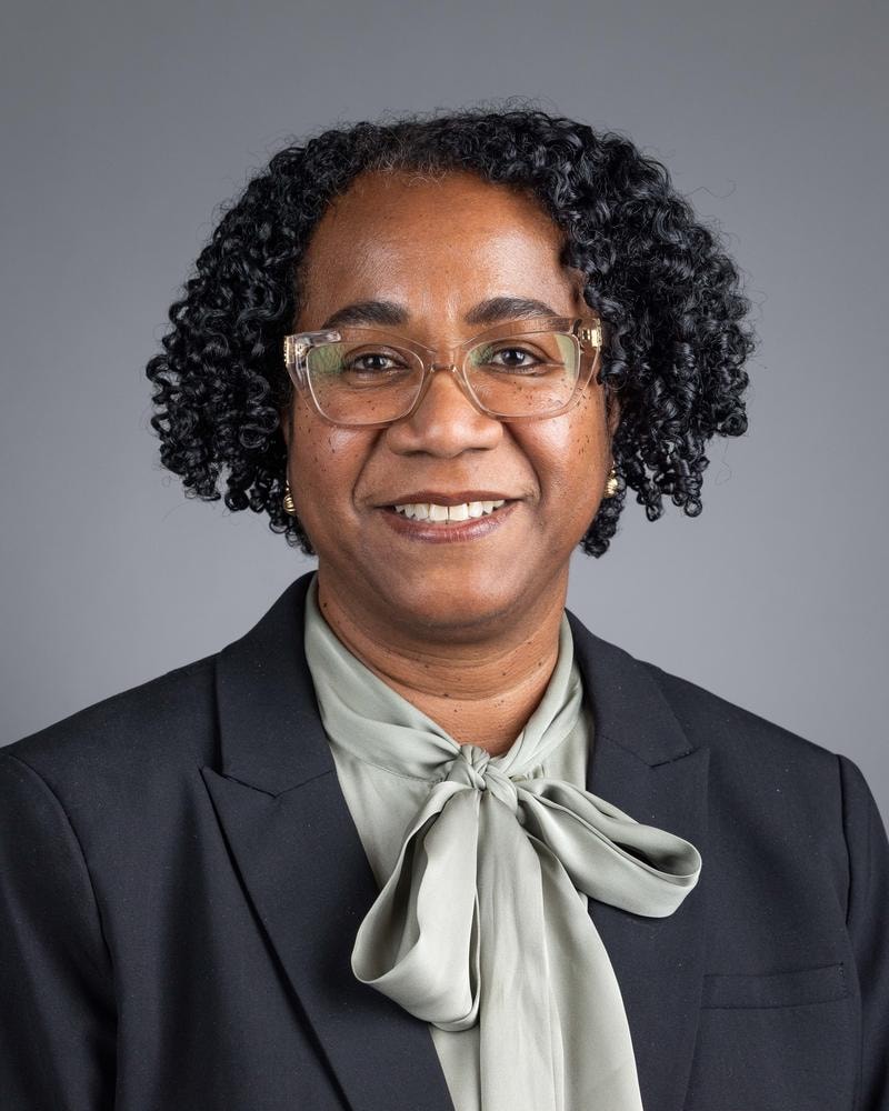 Executive headshot of a smiling woman in glasses and a bow blouse on a grey backdrop — corporate headshot by Michael Pegram